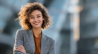 Happy young mixed race businesswoman with curly hair smiling confidently in gray blazer against blurred urban background for corporate marketing materials.