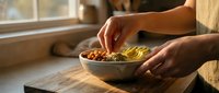 Woman hands garnishing healthy bowl with avocado slices and nuts on wooden cutting board in bright kitchen for nutritious meal preparation.