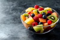 Fresh mixed fruit salad with berries, kiwi, mango and pineapple chunks in glass bowl on dark background, healthy breakfast or dessert serving.