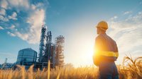 Male industrial worker in safety gear observing oil refinery plant at sunset, standing in golden wheat field with dramatic sky and sun rays creating silhouette effect.