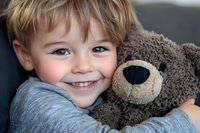 Cheerful young boy hugging soft brown teddy bear, showing bright genuine smile and blue eyes. Close-up portrait capturing innocent childhood joy and comfort with plush toy.