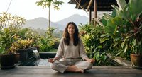 Young Asian woman meditating in lotus position on wooden deck surrounded by tropical plants with mountain landscape background during peaceful morning.