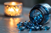 Blue medicine capsules spilled from glass bottle with warm candlelight bokeh in background, creating moody healthcare composition on wooden surface.