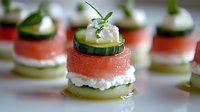 Layered appetizer bites with fresh cucumber, smoked salmon, cream cheese and herbs arranged on white plate. Elegant catering and party food presentation.
