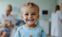 Smiling young boy in hospital gown with teddy bear pattern, showing resilience during medical treatment with healthcare professionals in background.