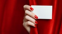 Female hand with red manicure holding blank white card against vibrant red fabric background, creating elegant contrast for business or holiday message.