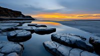 Dramatic coastal sunset over rocky beach with large fractured stones in foreground, reflecting golden light on calm water against moody blue twilight sky.