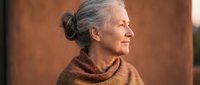 Senior woman with gray hair in bun, wearing rust-colored turtleneck sweater, shown in profile against warm brown background. Suitable for aging, lifestyle, and wellness content.