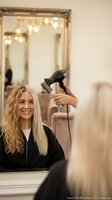 Young blonde woman getting professional hair styling with blow dryer at luxury salon, hairstylist working on client's hair in elegant mirror reflection.