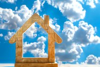 Wooden house outline cutout against bright blue sky with white clouds symbolizing home ownership dreams and real estate investment opportunities.