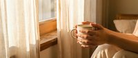 Woman holding a warm ceramic mug near a sunlit window with sheer curtains, cozy morning atmosphere with soft golden light.
