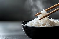 Wooden chopsticks lifting steaming white rice from black bowl against dark background, suitable for food, cuisine, and Asian cooking content.