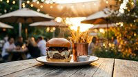 Gourmet burger with fries served on wooden table at outdoor restaurant patio during golden hour with string lights and blurred diners in background.