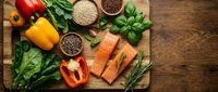 Fresh salmon fillet with colorful bell peppers, leafy greens, quinoa and herbs on wooden cutting board for healthy cooking and meal preparation.