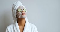 Young woman in white bathrobe and towel enjoying spa treatment with cucumber eye mask and facial sheet mask for skincare routine and relaxation.