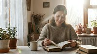 Young Asian woman in cozy sweater reading book at home with coffee, surrounded by houseplants and warm natural light streaming through window.