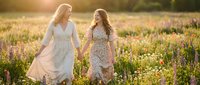 Two young women holding hands walking through wildflower meadow at golden hour sunset wearing flowing dresses in natural outdoor setting for friendship concepts.