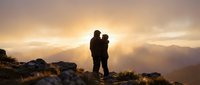 Silhouette of romantic couple embracing on mountain peak during golden sunset with dramatic sky and layered mountain ranges in background for travel and adventure concepts.