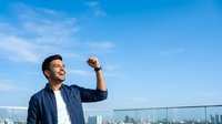 Man in casual navy shirt and white undershirt flexing arm muscle outdoors against blue sky and city skyline background, showing strength and confidence for fitness, wellness, and motivation concepts.