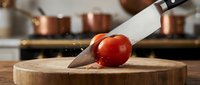 Knife cutting fresh red tomato on wooden chopping board in kitchen with copper cookware in background.