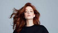 Young redhead woman with flowing hair against light background, wearing black top, looking up with confident expression. Natural makeup and dynamic hair movement.