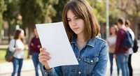 Young brunette woman in denim jacket reading document outdoors on campus with blurred students in background for education and academic success concepts.