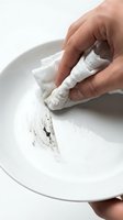 Hand cleaning white ceramic plate with cloth removing food residue on neutral background for kitchen and household cleaning concepts.