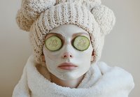 Young woman wearing white knit hat and bathrobe with facial clay mask and cucumber slices on eyes during spa skincare treatment and relaxation routine.