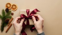 Hands tying burgundy ribbon bow on gift box with dried oranges, pine cones, cinnamon sticks and evergreen branch for festive holiday wrapping preparation.