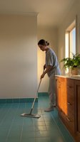 Woman in casual clothing mopping teal tile floor in bright hallway with wooden vanity and potted plant, domestic cleaning and household chores.