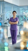 Young Asian female healthcare professional wearing purple scrubs standing confidently with crossed arms in modern medical facility with glass partitions and laboratory equipment visible in background.