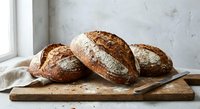 Three rustic artisan bread loaves with crusty exterior on wooden cutting board near window, perfect for bakery marketing and food photography projects.