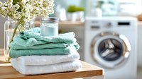 Fresh mint green and white folded towels stacked on wooden surface with glass jar and elderflower bouquet, washing machine in background for laundry and bathroom decor.