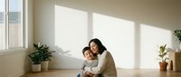 Young Asian mother embracing toddler son in sunlit minimalist living room with houseplants creating warm family bonding moment for parenting content.