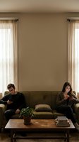 Young couple sitting on opposite ends of couch using smartphones in modern living room with natural lighting and neutral decor.