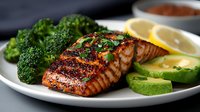 Grilled salmon fillet with fresh broccoli, avocado slices and lemon on white plate. Perfectly seared fish with herbs and spices shows golden-brown crust and moist center.