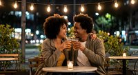 Happy African American couple enjoying romantic date night with drinks and string lights creating warm ambiance at outdoor restaurant terrace.