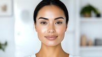 Young Asian woman with natural makeup and glowing skin smiling confidently at camera against bright minimal interior background with decorative shelves.