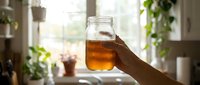 Hand holding a glass jar filled with cold brew tea or iced beverage against a bright window with hanging plants and natural sunlight streaming through, perfect for beverage, wellness, and lifestyle marketing.