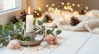 Cozy winter home decor with burning candle, eucalyptus, pine cones, fairy lights and knitted blanket creating warm festive atmosphere for holiday season.
