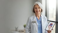 Smiling mature Caucasian female doctor in white coat holding tablet displaying heart anatomy diagram in modern medical office for healthcare education.