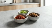 Three ceramic bowls filled with colorful spices and herbs arranged on marble countertop in modern kitchen setting for culinary cooking and food preparation.