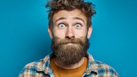 Surprised bearded man with wide eyes and raised eyebrows expressing shock or amazement against bright blue background for advertising and marketing campaigns.