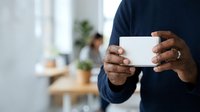 Cropped view of male professional with dark skin tone wearing navy blue shirt holding blank white business card in modern office environment with colleagues and plants in background.