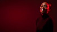 Young African American woman laughing joyfully with red neon lighting against dark background for creative portrait photography and advertising campaigns.