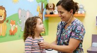 Female pediatric nurse examining young girl with stethoscope in colorful children's medical office with animal wall decorations and toys.