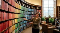 Middle-aged Caucasian woman librarian standing in curved library with colorful organized books by rainbow spectrum, natural window light, cozy reading area.