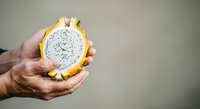 Close-up of hand holding halved yellow dragon fruit with white flesh and black seeds against neutral background, showcasing tropical fruit texture and freshness.