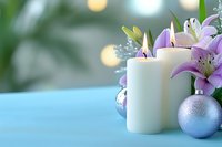 Glowing white candles with pink lilies and silver Christmas ornaments on blue surface against blurred bokeh background create festive holiday atmosphere.