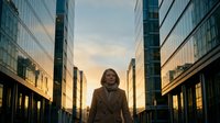 Woman in business attire standing confidently in urban canyon of modern skyscrapers during golden hour sunset, suitable for corporate leadership and professional success concepts.